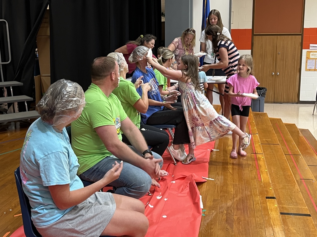 A group of adults sit in a line of chairs as part of a pie-in-the face event. A child pushes a pie into one of their faces. 