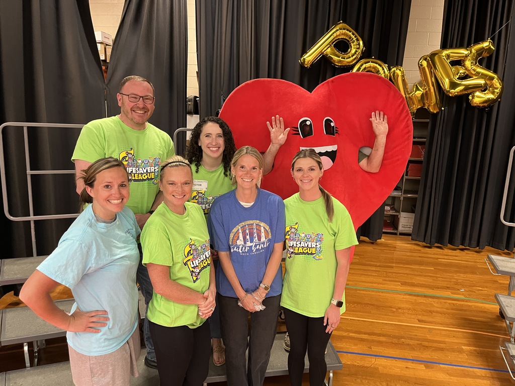 A group of school employees and a guest pose for a photo with a person in a heart costume.