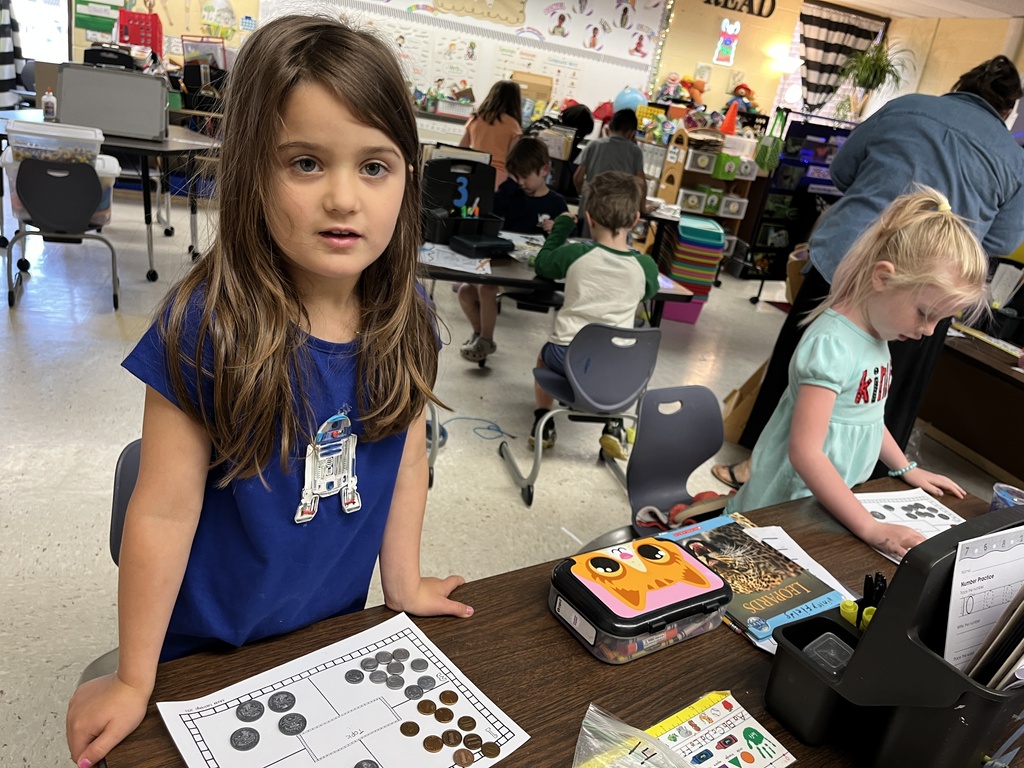 Kindergartners learns about the value of money by working with replica coins. A teacher helps one of the students.