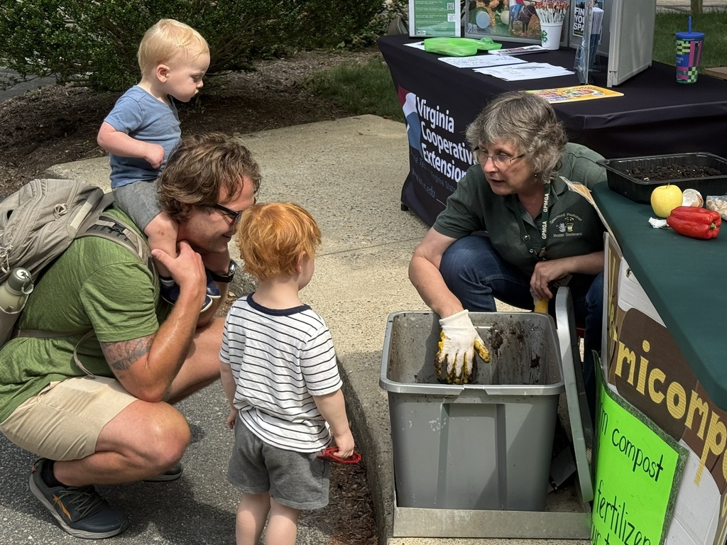 An adult and two children listen to a woman talk about composting at a summer reading program kickoff event.
