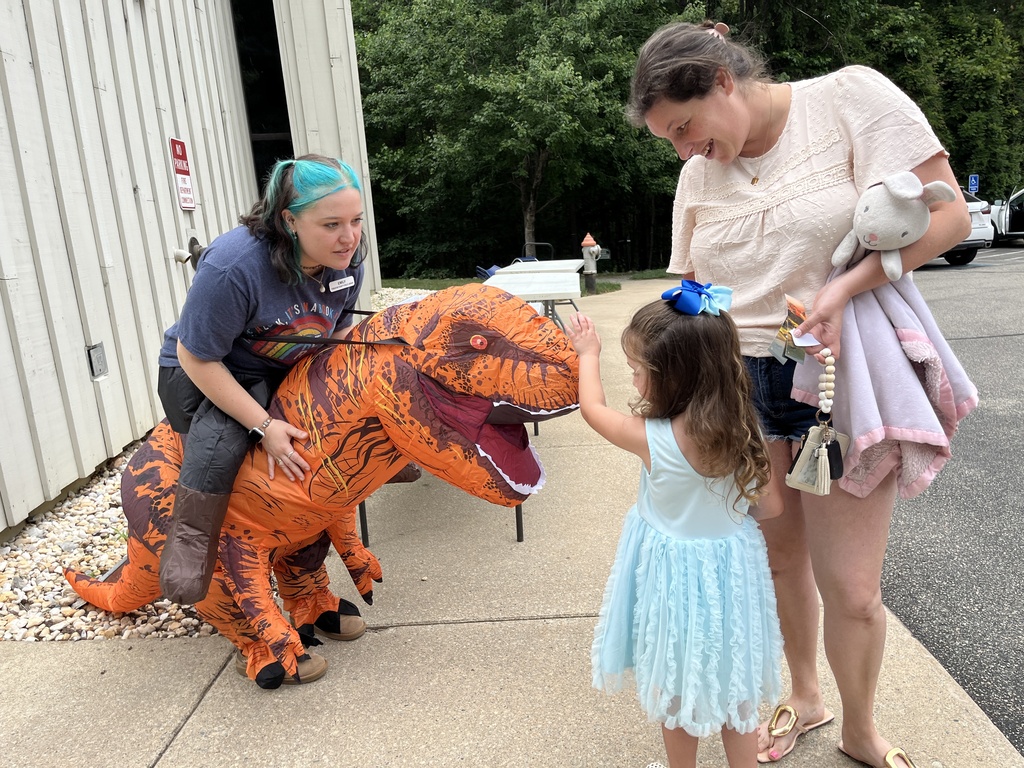An adult and child talk to a woman in a dinosaur costume at a summer reading program kickoff event.