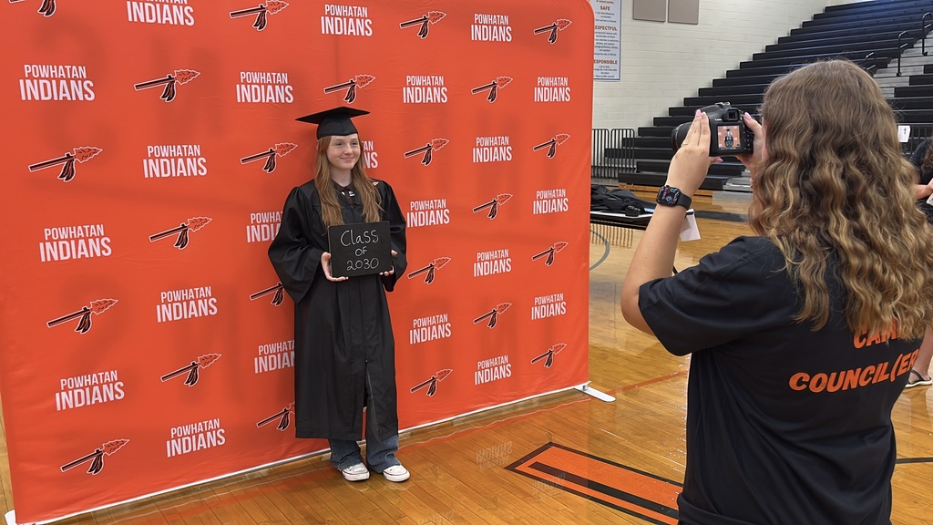 A student takes a photo of a freshman wearing graduation garb and holding a Class of 2030 sign on the first day of school.