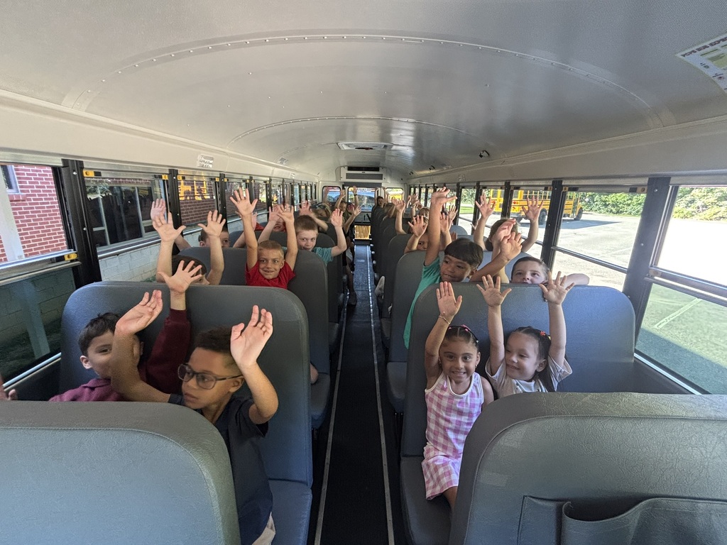 A group of first graders listen to a bus driver on a school bus talk about bus safety. All of their hands are raised.