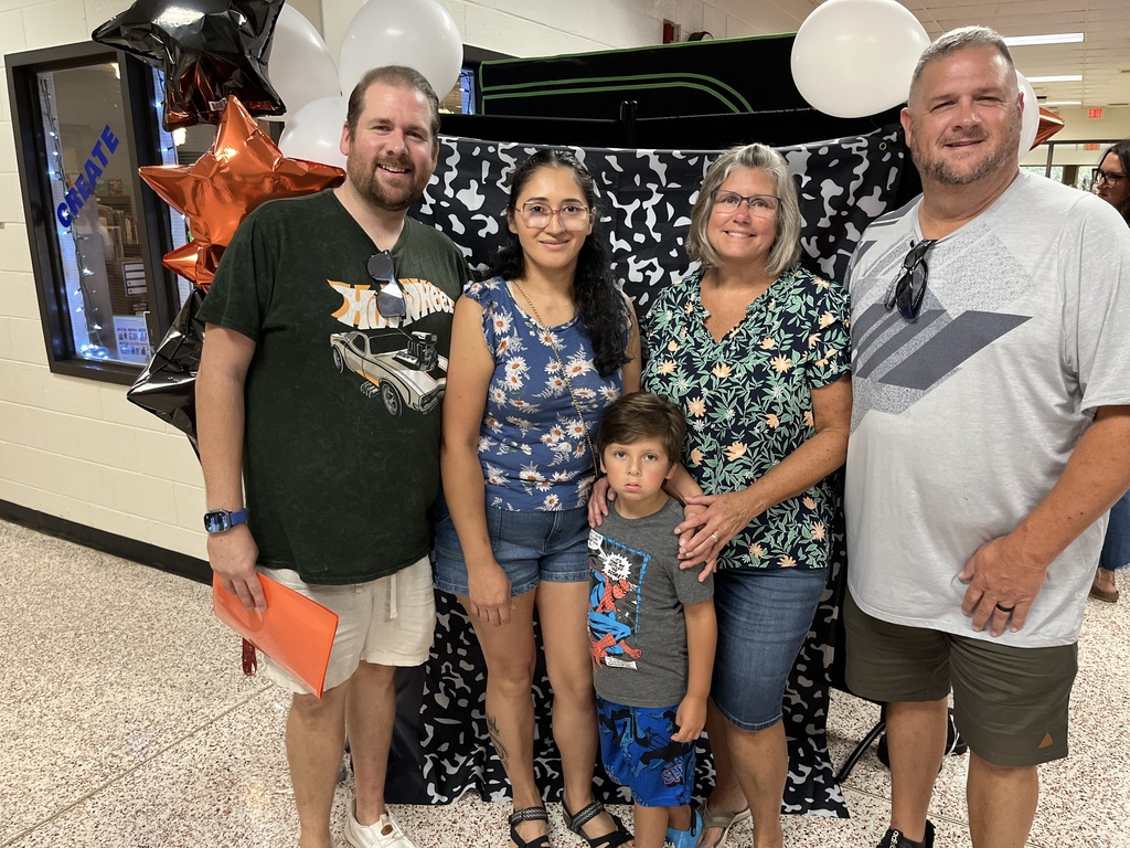 Four adults and a child pose for a photo in front of a photo display at an elementary open house. 