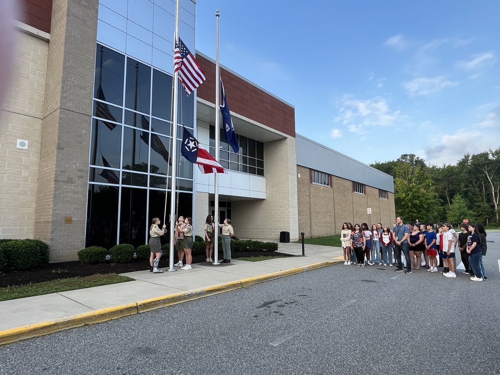 Scouting America members raise the flag during a 9/11 ceremony held in front of a middle school. A student chorus group watches. 