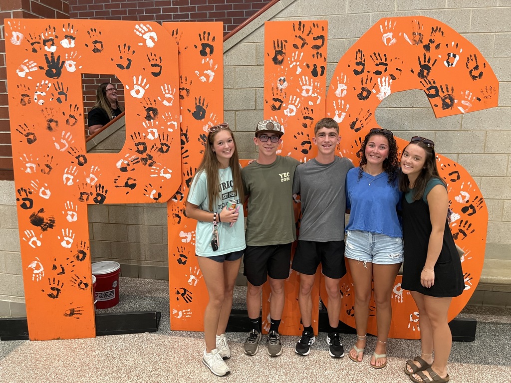 Five teenagers pose for a photo in front of a large orange PHS backdrop at a high school open house event.