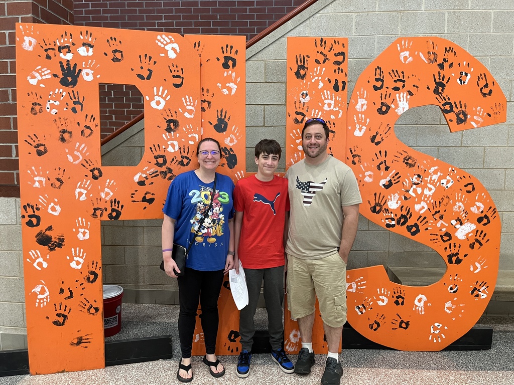 Two adults and a child pose for a photo in front of a large orange PHS backdrop at a high school open house event.