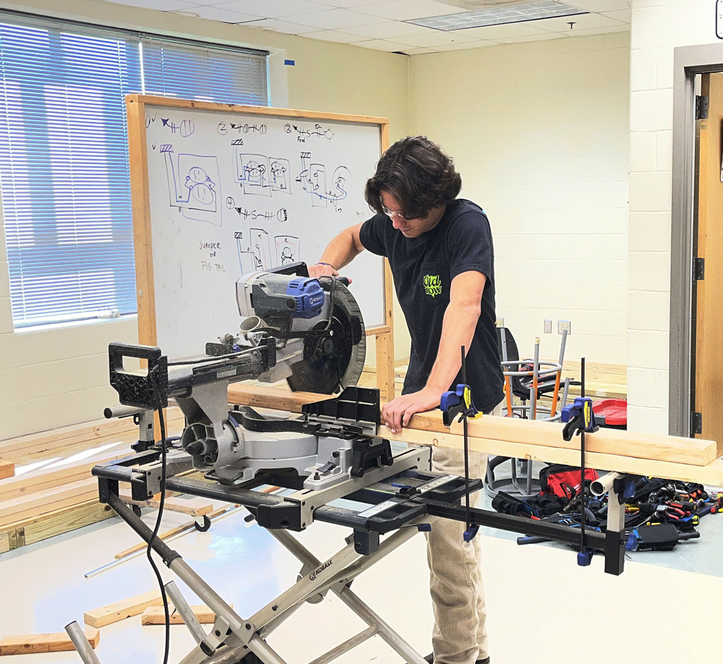 A student uses a saw to cut woord that will be used to build electricity booths. 
