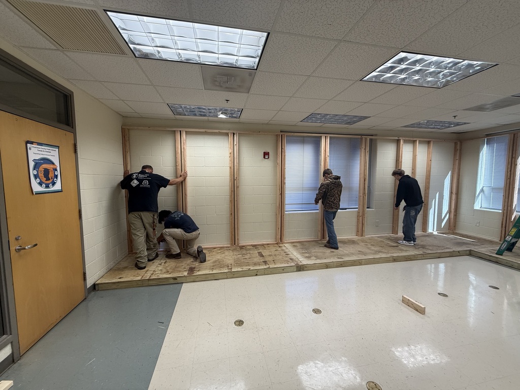 Building trades students measure a wood floor to build electricity booths. A teacher supervises. 