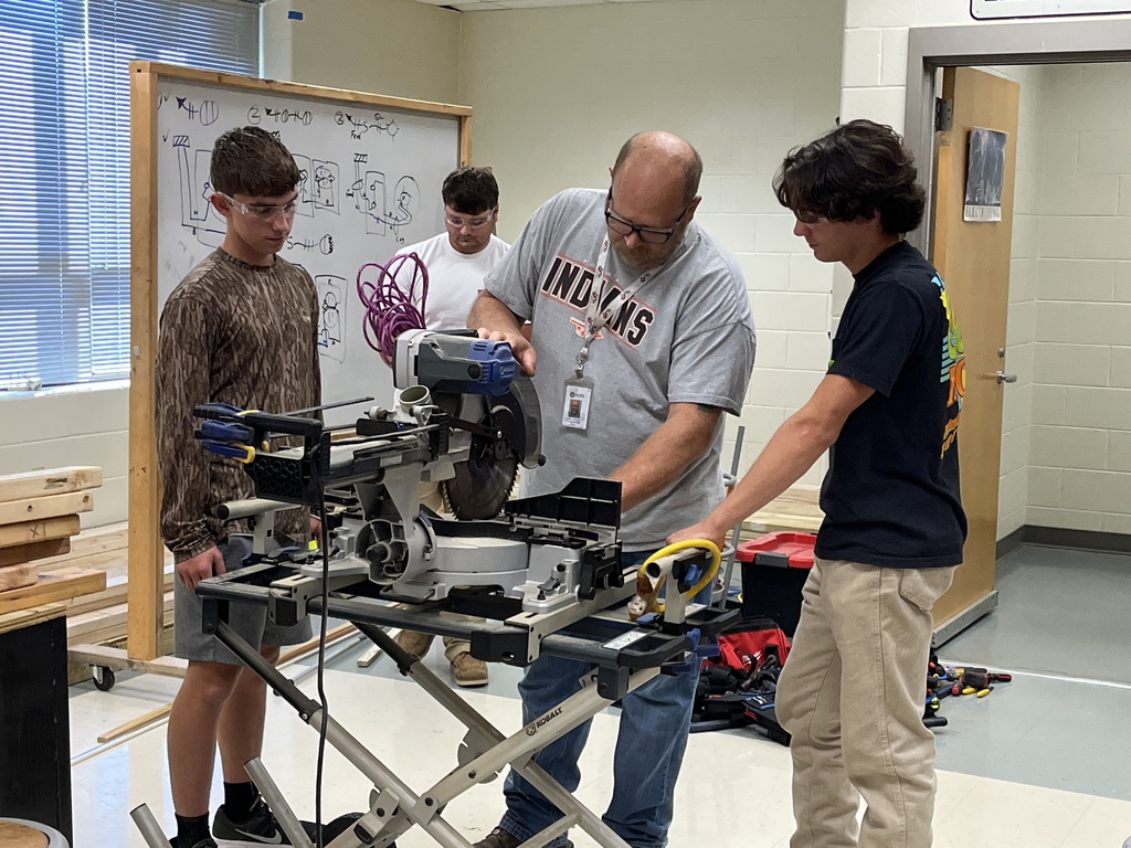 A teacher demonstrates how to use a saw to building trades students building electricity booths. 
