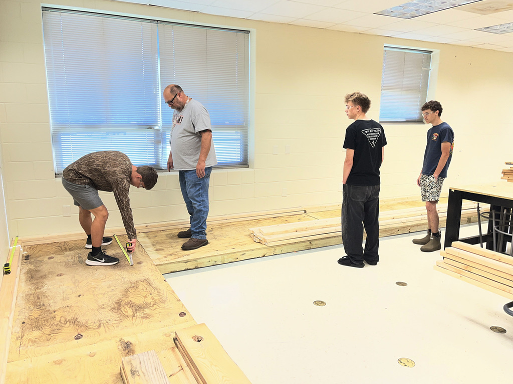 Building trades students measure a wood floor to build electricity booths. A teacher supervises. 