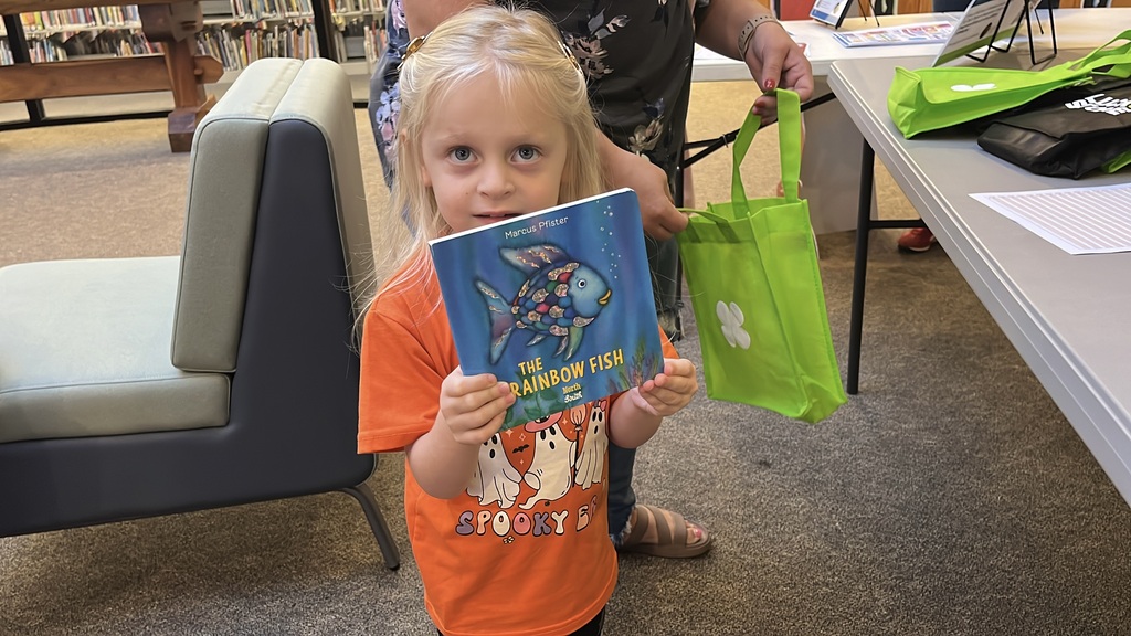 A small child holds up a copy of the book, The Rainbow Fish.