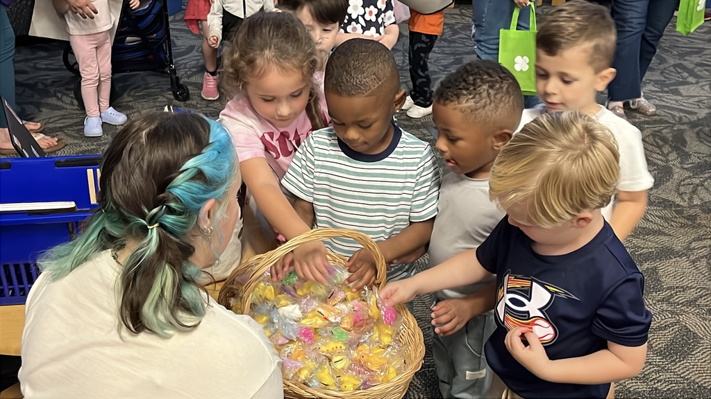 Children take a small prize from a basket being held by a librarian at a preschool literacy event at a library.