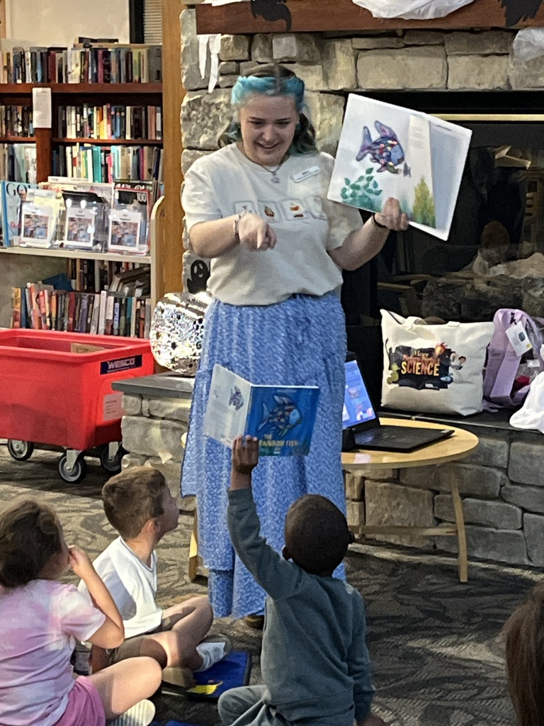 Children sit and listen to a librarian reading a story during a preschool literacy event at a library. Adults watch. 