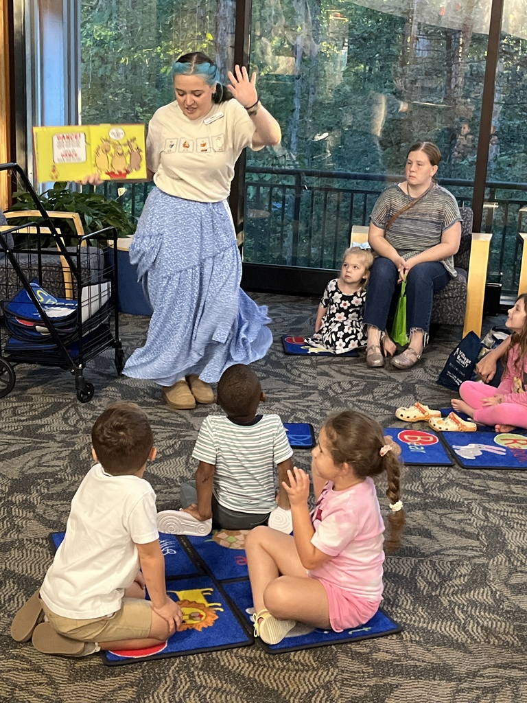 Children sit and listen to a librarian reading a story during a preschool literacy event at a library. Adults watch. 
