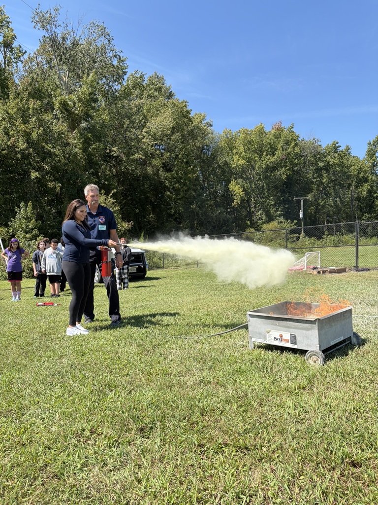 A firefighter lets a student use a fire extinguisher to put out a controlled fire to learn about fire safety. Students and adults standing nearby watch.