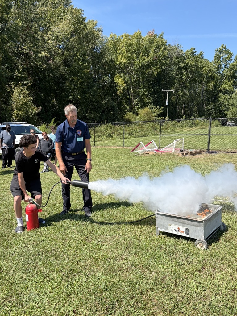 A firefighter lets a student use a fire extinguisher to put out a controlled fire to learn about fire safety. Students and adults standing nearby watch.