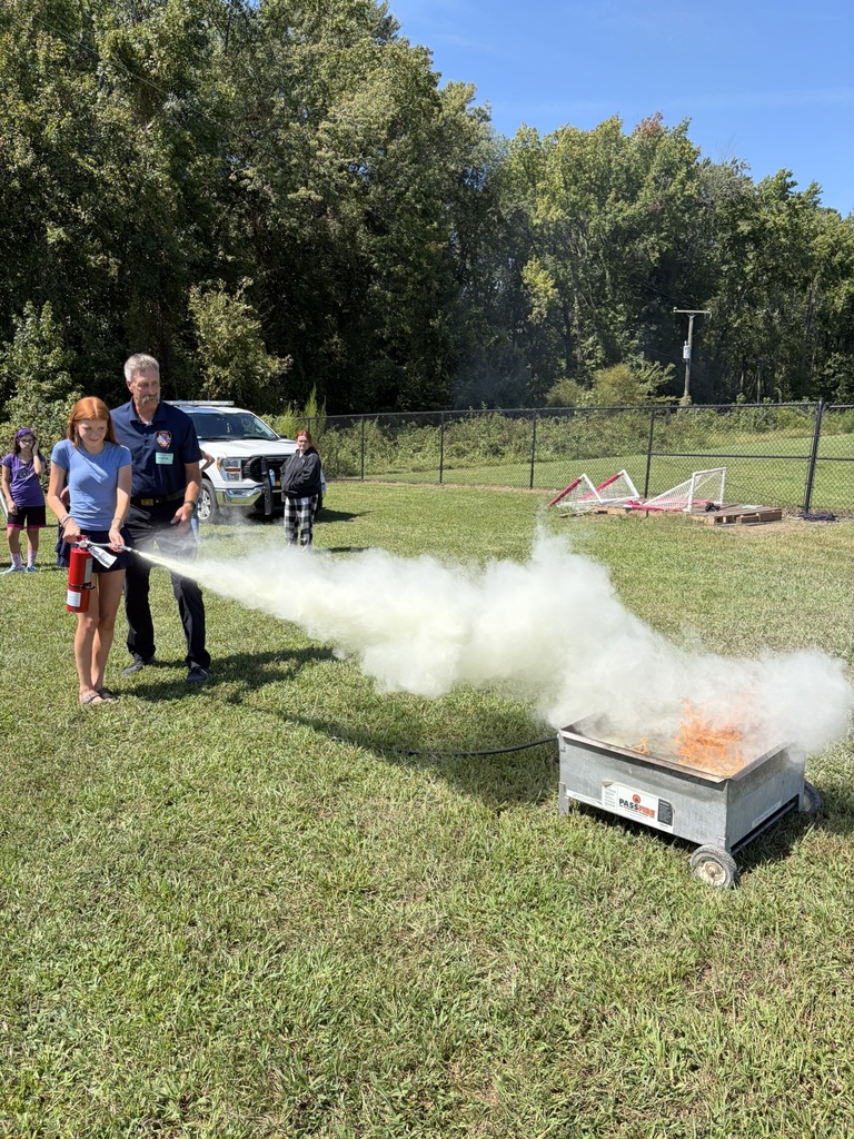 A firefighter lets a student use a fire extinguisher to put out a controlled fire to learn about fire safety. Students and adults standing nearby watch.