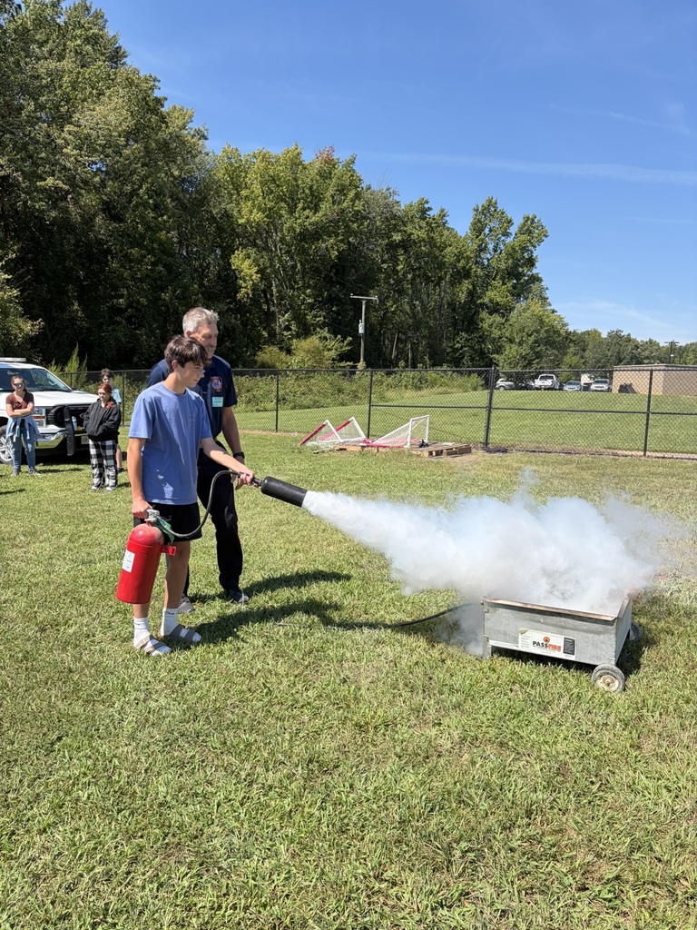 A firefighter lets a student use a fire extinguisher to put out a controlled fire to learn about fire safety. Students and adults standing nearby watch.