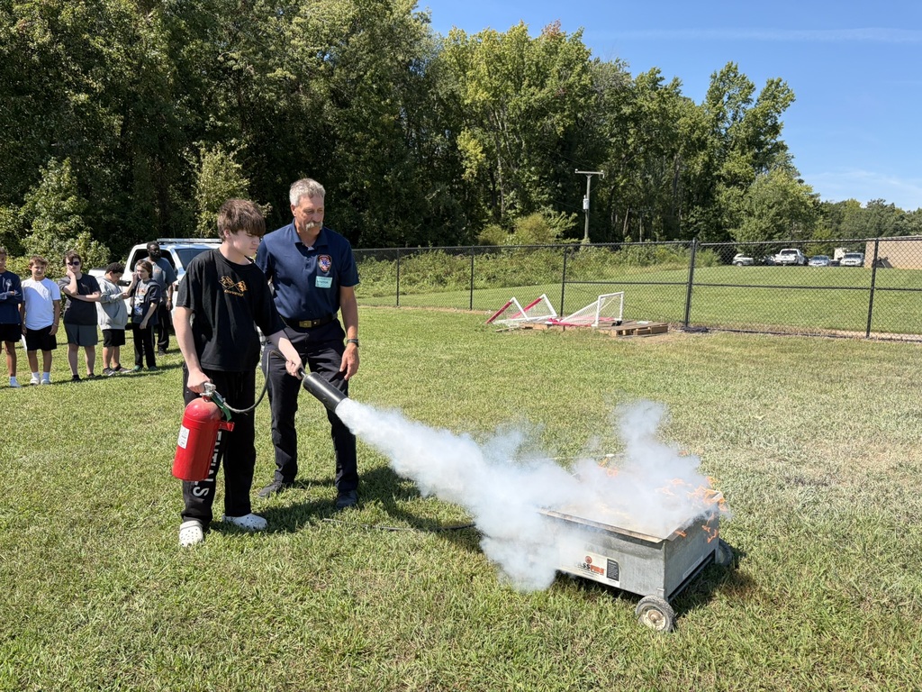 A firefighter lets a student use a fire extinguisher to put out a controlled fire to learn about fire safety. Students and adults standing nearby watch.