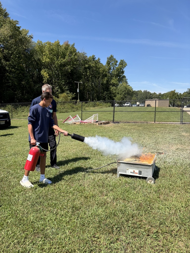 A firefighter lets a student use a fire extinguisher to put out a controlled fire to learn about fire safety. Students and adults standing nearby watch.