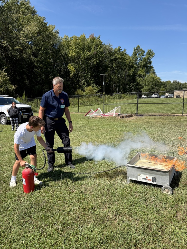 A firefighter lets a student use a fire extinguisher to put out a controlled fire to learn about fire safety. Students and adults standing nearby watch.