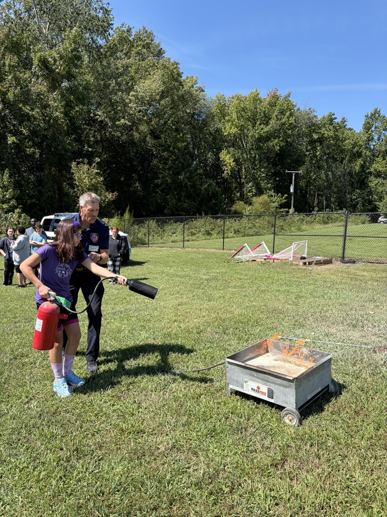 A firefighter lets a student use a fire extinguisher to put out a controlled fire to learn about fire safety. Students and adults standing nearby watch.