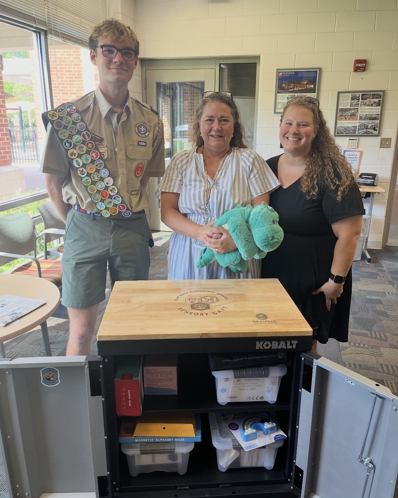 Two adults and a teen in an Eagle Scout uniform pose for a photo in front of a sensory cart the Scout donated.