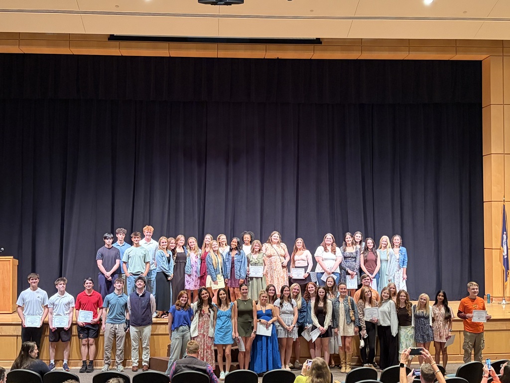 A large group of students pose for a photo in two rows on and in front of an auditorium stage.