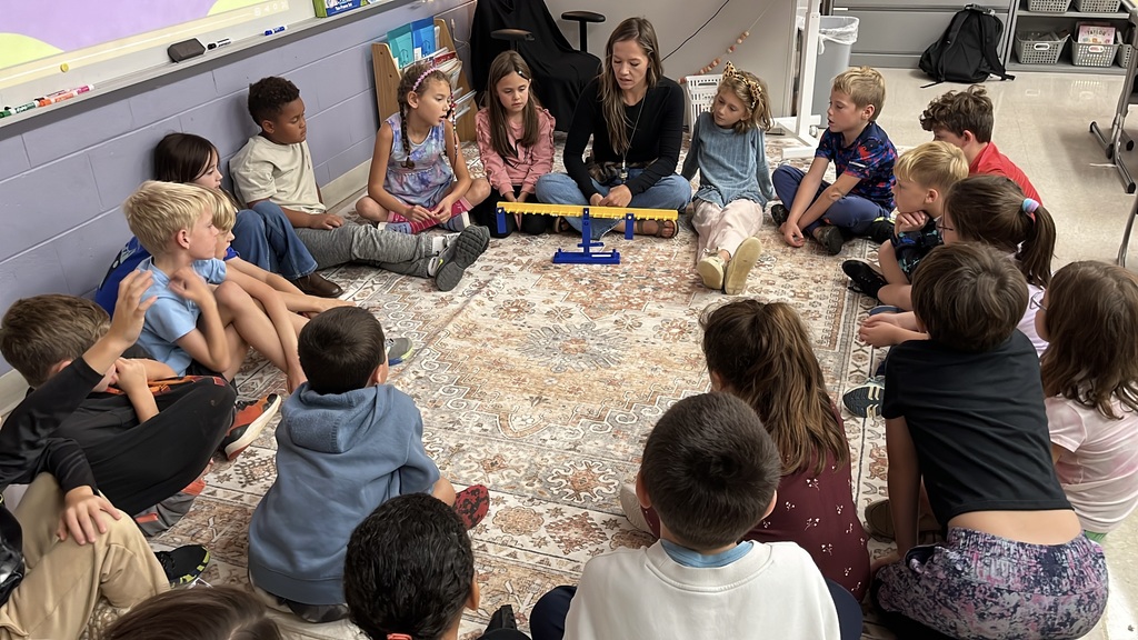 Students sit in a circle listening to their teacher explain how a balance works as they explore equality and inequality.