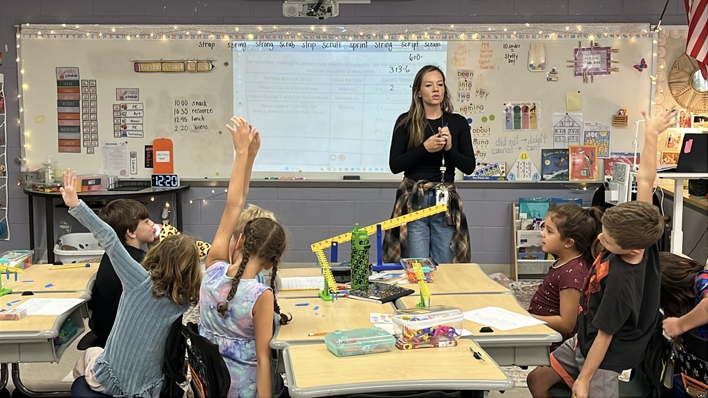 Students  listen to their teacher explain how a balance works as they explore equality and inequality. Some students make a tilted balance symbol with their bodies in alignment with the lesson.
