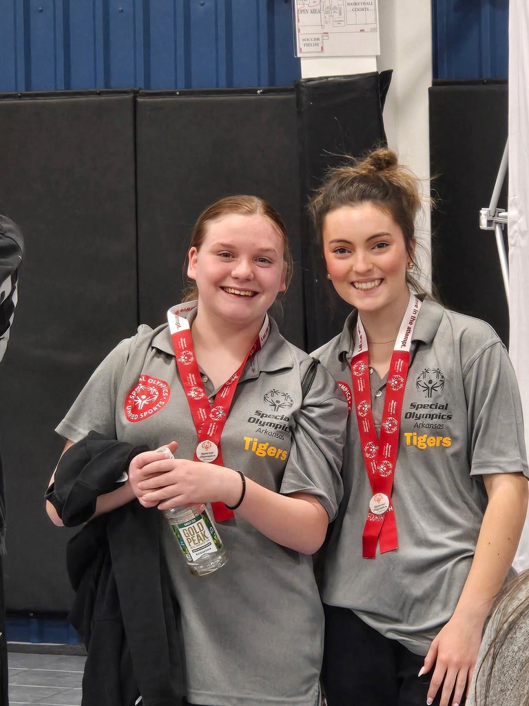 2 students standing together and smiling up at the special olympics award platform after receiving their medals
