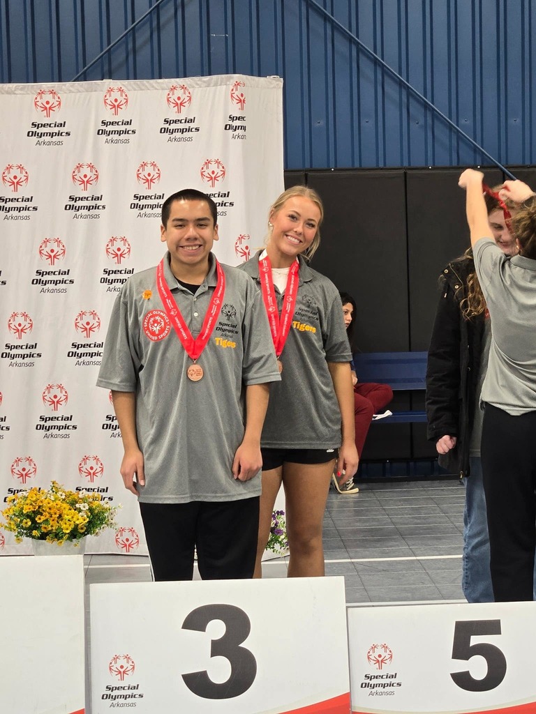 2 students standing together and smiling up at the special olympics award platform after receiving their medals