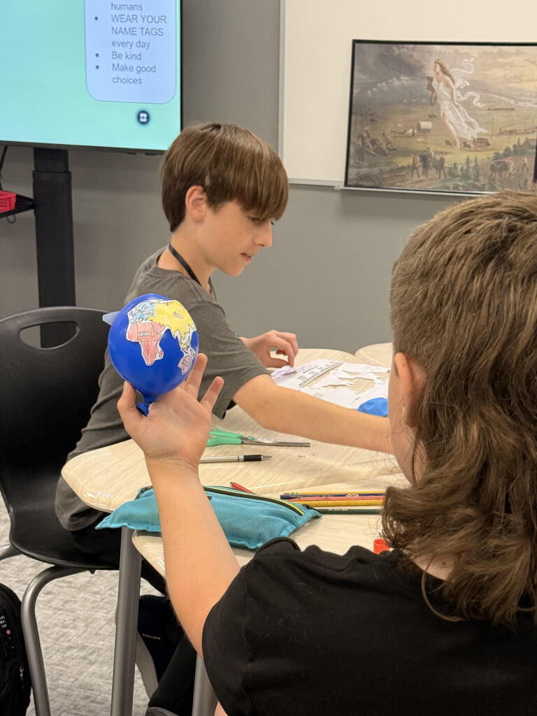 Two students sit together at a classroom table, attaching colored map pieces to blue balloons as part of a geography activity. Colored pencils, scissors, glue, and worksheets are on the table.