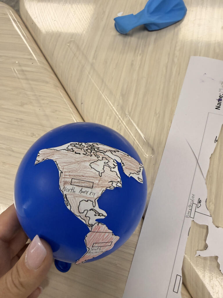 A close-up of a blue balloon decorated with hand-colored paper cutouts representing North and South America. The continents are labeled, with North America colored brown and South America colored red. A student holds the balloon beside a worksheet with additional map pieces.