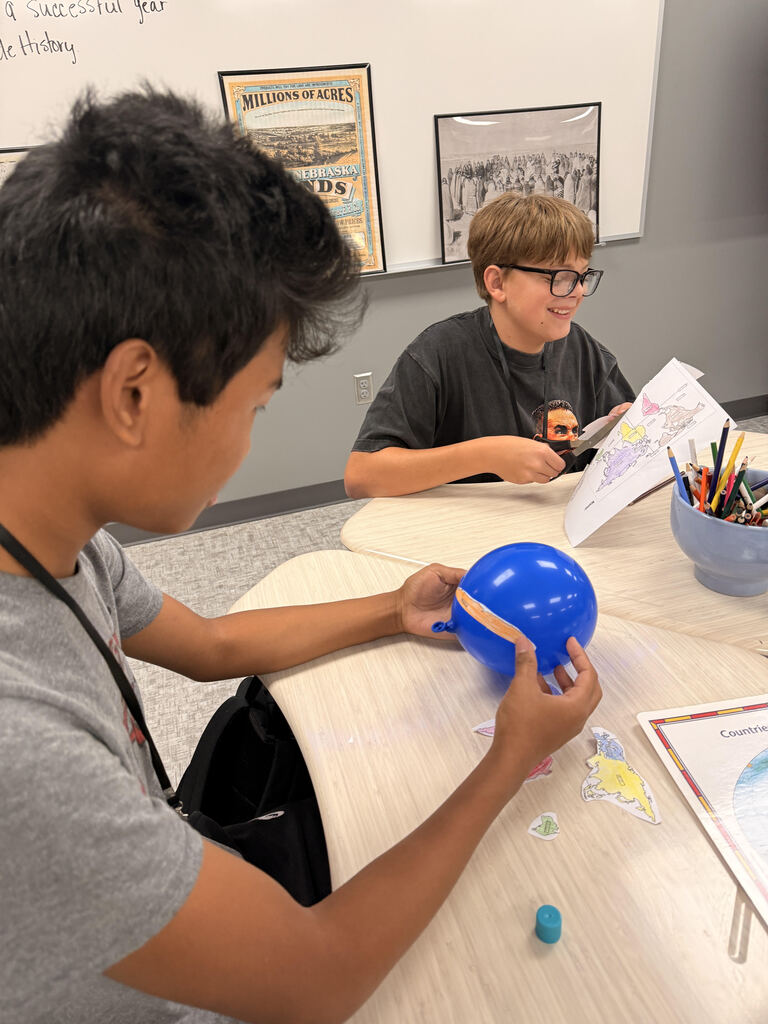 A student cuts out colored map pieces at a classroom table while another student works nearby. A blue balloon, map pieces, colored pencils, and other supplies are spread across the table.