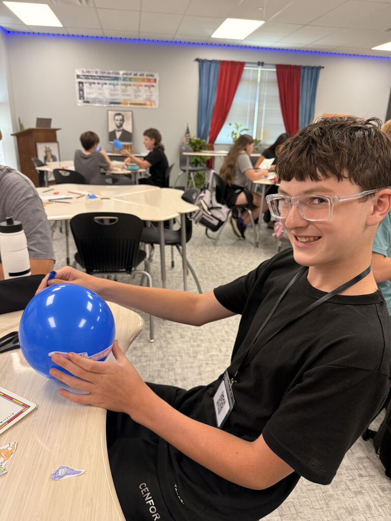 A student smiles while holding a blue balloon decorated with colored paper map pieces. Other students work on the same geography activity at tables in the classroom behind him.
