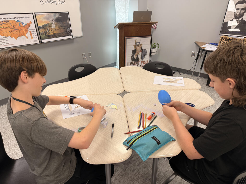 Two students work together on a geography activity at a classroom table. One student holds up a blue balloon decorated with colored paper continents while the other works with map pieces and a worksheet.