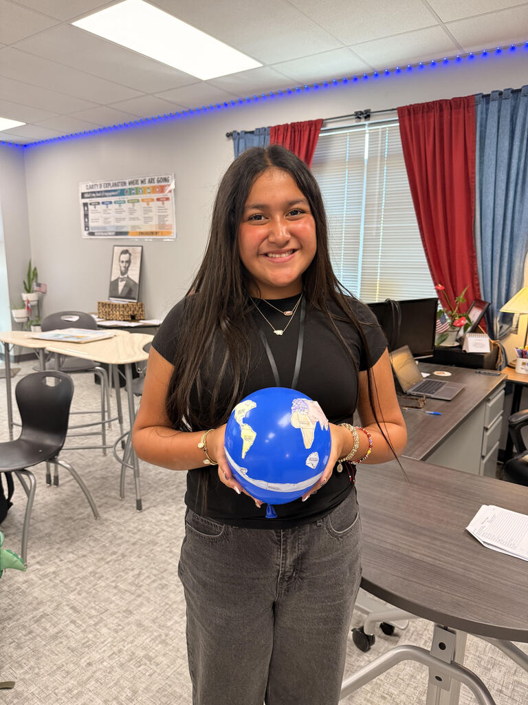 A smiling student stands in a classroom holding a blue balloon decorated with colored paper continents representing the world. Desks, computers, and history-themed classroom displays are visible in the background.