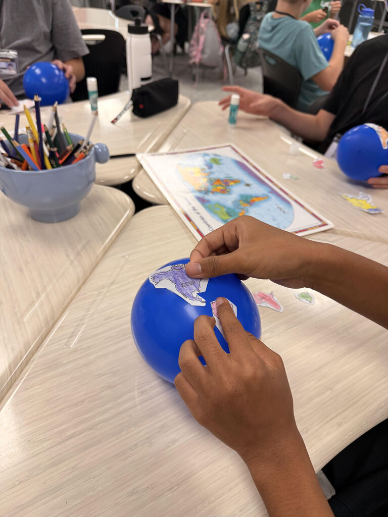 Students work together at classroom tables on a geography activity. One student places a colored paper map piece onto a small blue balloon representing Earth, while classmates work with balloons, maps, colored pencils, and other craft supplies nearby.