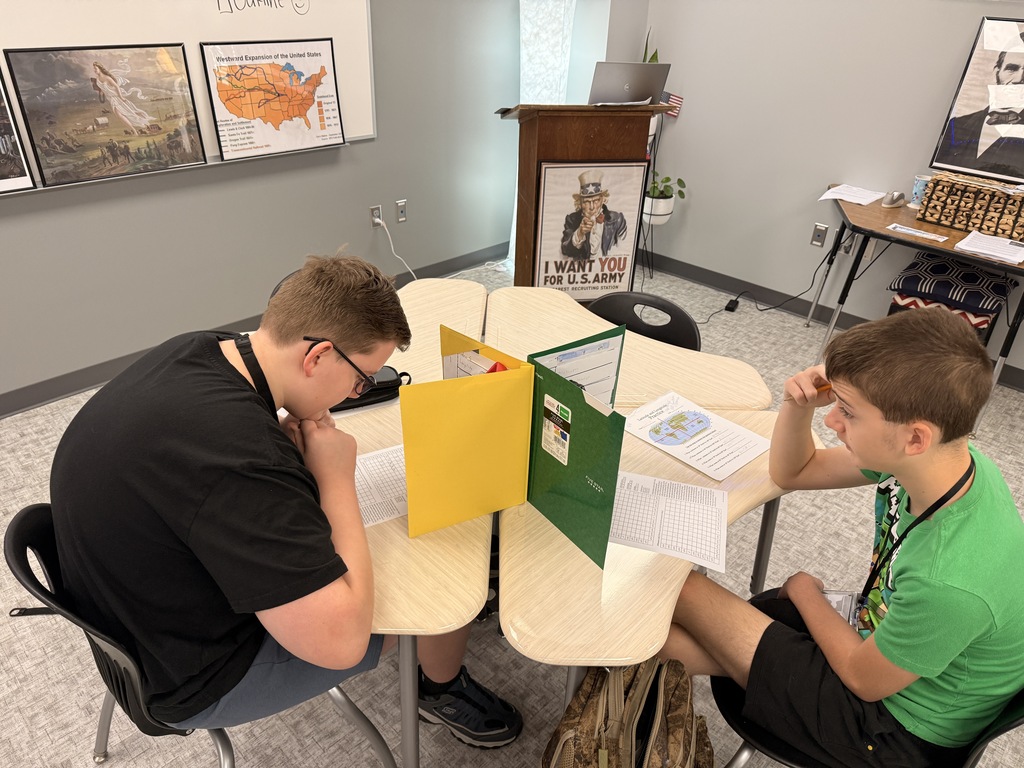 Two students battleship at their desks in a history classroom with maps, historical artwork, and a U.S. Army poster is visible in the background.