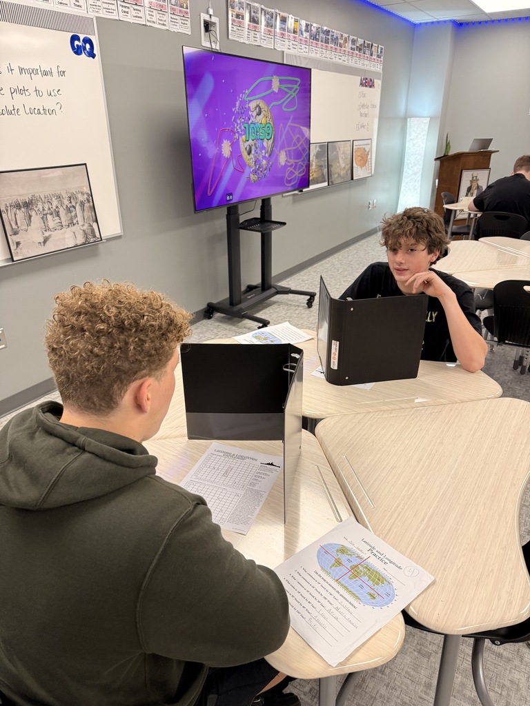 student uses a pencil to complete a latitude and longitude battleship activity at a classroom desk. A black binder stands upright as a privacy divider, with a geography worksheet visible on the table.