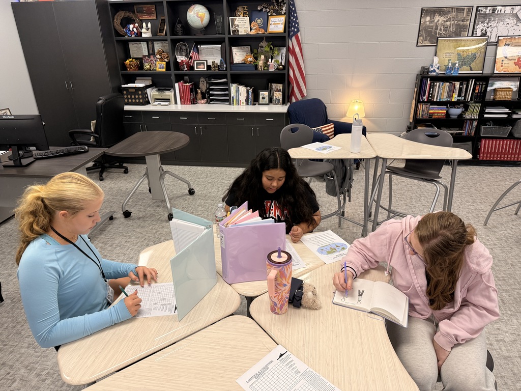 Three students work together at classroom tables on geography assignments. Two students write on worksheets while another works in a notebook, with colorful binder dividers, school supplies, books, and history displays visible in the classroom.