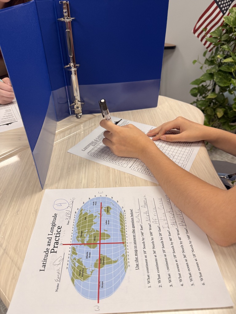 Students complete a latitude and longitude game  at a classroom table. One student writes on a worksheet while using a blue binder as a privacy divider, with a world map and coordinates visible on the page.