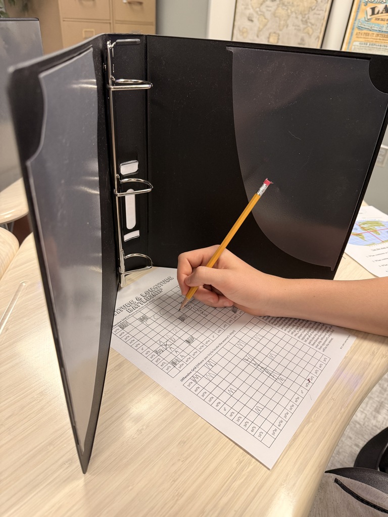 A student uses a pencil to complete a latitude and longitude battleship activity at a classroom desk. A black binder stands upright as a privacy divider, with a geography worksheet visible on the table.