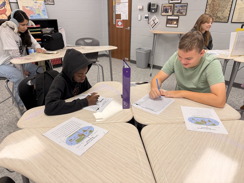 Students work on latitude and longitude game at desks throughout a classroom. One student writes on a worksheet while another reads from a worksheet, with maps and geography materials visible around the room.