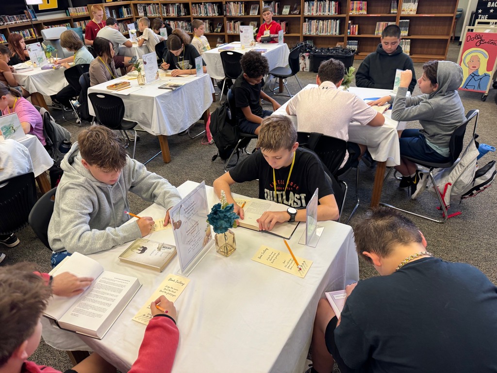 Students are seated in the library at tables while they read and complete a book tasting form.