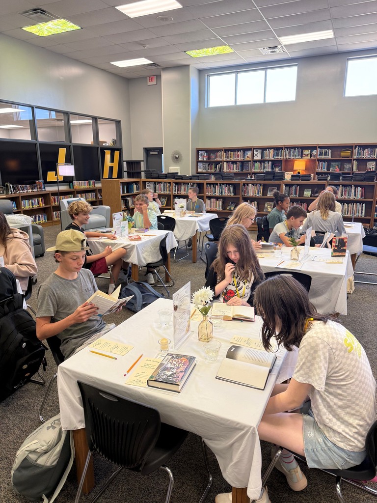 Students in the library are browsing books at tables with tablecloths, flowers and book tasting forms.