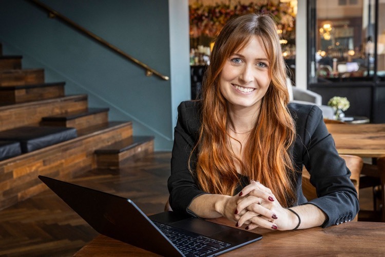 woman sitting with open computer smiles at camera. she is in a modern office meeting space designed with wooden floors and wooden furniture.