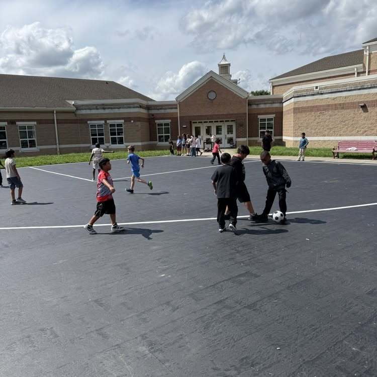 Stewart Elementary students playing on a brand-new painted playground with the school in the background. Three of the students are kicking around a soccer bowl with another child holding another ball.